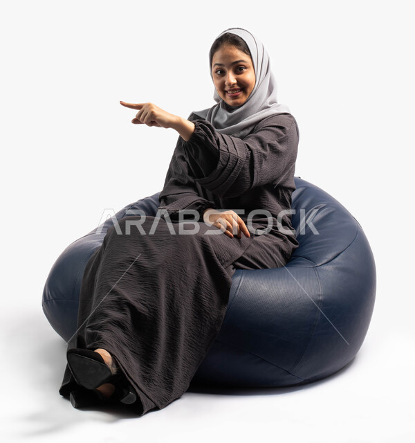 إيماءات وتعابير وجه تدل على رؤية شيء جميل , بورتريه لإمرأة عربية سعودية خليجية تجلس على كرسي مريح و ترفع يدها للأعلى, الإشارة الى شيء ما , خلفية بيضاء