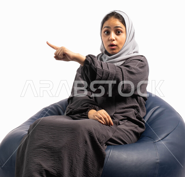 إيماءات وتعابير وجه تدل على الدهشة والتفاجؤ , بورتريه لإمرأة عربية سعودية خليجية تجلس على كرسي مريح و ترفع يدها للأعلى, الإشارة الى شيء ما , خلفية بيضاء