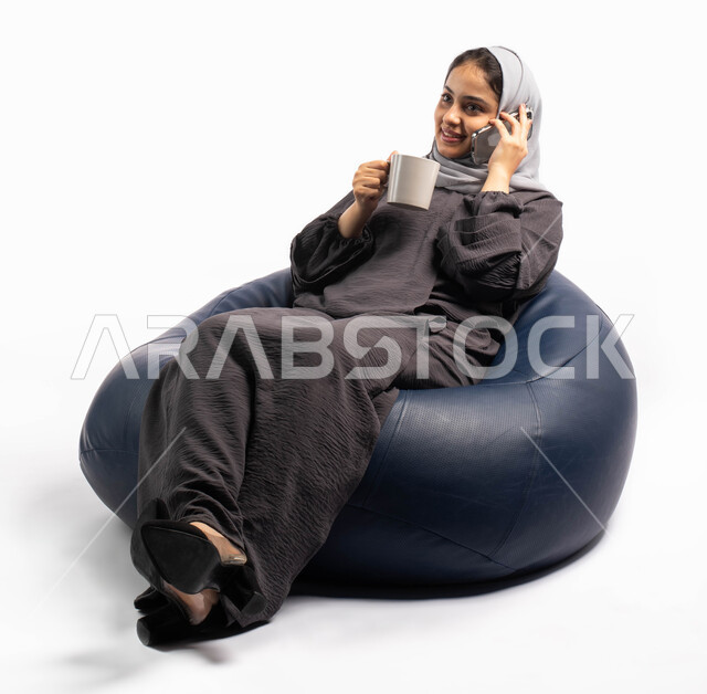 Communicating with family and friends, gestures indicating happiness and enjoying making a phone call, a portrait of a Saudi Arabian Gulf woman sitting on a comfortable chair holding a cup of coffee and talking on a mobile phone, using electronic programs