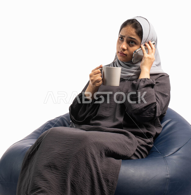 استخدام البرامج والتطبيقات الالكترونية، التواصل مع العائلة والأصدقاء بإجراء مكالمة هاتفية، ايماءات تدل على الضجر والحزن , بورتريه لإمرأة عربية خليجية سعودية تجلس على كرسي مريح تحمل بيدها فنجان القهوة وتتحدث عبر الهاتف المحمول، خلفية بيضاء
