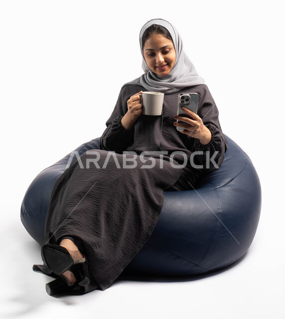تصفح المواقع الالكترونية والتواصل الاجتماعي، ايماءات تدل على السعادة والاستمتاع بإجراء مكالمة فيديو, بورتريه لإمرأة عربية خليجية سعودية تجلس على كرسي مريح تحمل بيدها فنجان القهوة وتستخدم الهاتف المحمول ، استخدام البرامج والتطبيقات الالكترونية، خلفية بيضاء