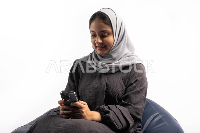 Online shopping, browsing websites and social networking, using electronic programs and applications, having fun, a portrait of a Saudi Gulf Arab woman sitting on a comfortable chair browsing the mobile phone, white background