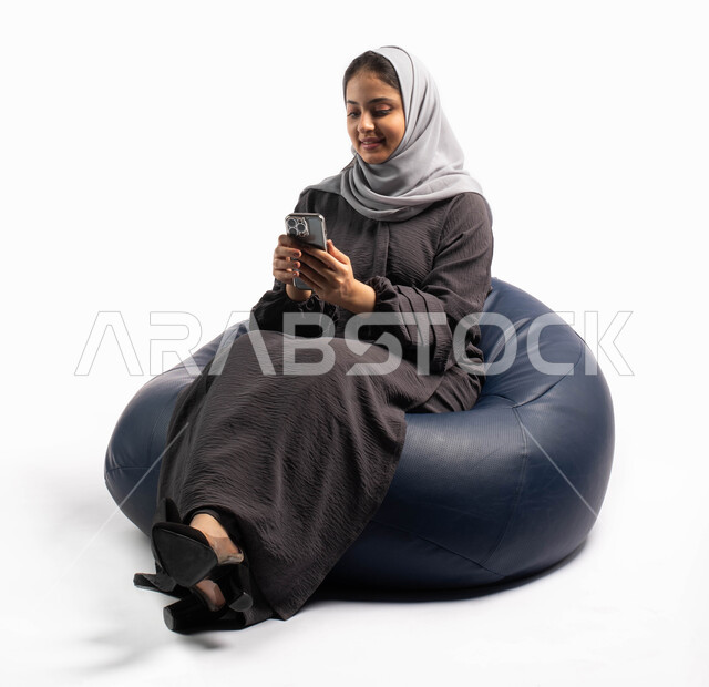 Browsing websites and social communication, using electronic programs and applications, having fun, a portrait of a Saudi Arabian Gulf woman sitting on a comfortable chair browsing a mobile phone, online shopping, white background