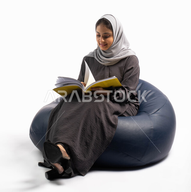 إيماءات تعبر عن الاستمتاع بقراءة الكتب, ثقافة التعلم الذاتي, إنجاز الواجبات اليومية, قضاء الوقت بالقراءة والتعلم والتثقف ، بورتريه لإمرأة عربية سعودية خليجية تجلس على كرسي مريح وتحمل بيديها كتاب، خلفية بيضاء