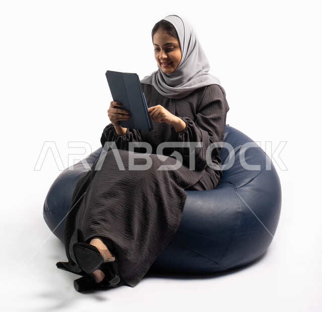 التواصل مع العائلة والأصدقاء عبر الجهاز اللوحي, إيماءات وجه ويدين تعبر عن الاستمتاع بتصفح الإنترنت وإنجاز المهام عن بعد، بورتريه لامرأة عربية سعودية خليجية تتجلس على كرسي مريح تستخدم جهاز التابلت ، التسوق الإلكتروني الآمن، خلفية بيضاء 