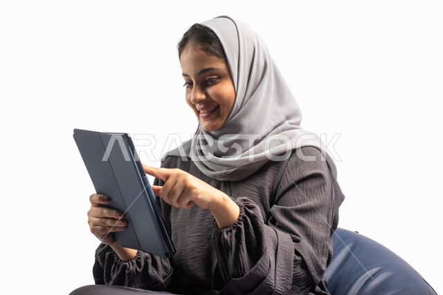 التواصل مع العائلة والأصدقاء عبر الجهاز اللوحي, إيماءات وجه ويدين تعبر عن الاستمتاع بتصفح الإنترنت وإنجاز المهام عن بعد، بورتريه لامرأة عربية سعودية خليجية تتجلس على كرسي مريح تستخدم جهاز التابلت ، التسوق الإلكتروني الآمن، خلفية بيضاء 