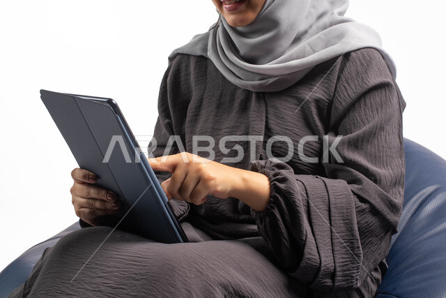 قضاء الوقت بتصفح الجهاز اللوحي, إستخدام اجهزة تقنية حديثة،  بورتريه لامرأة عربية سعودية خليجية تتجلس على كرسي مريح تستخدم جهاز التابلت ، التسوق الإلكتروني الآمن، تصفح الإنترنت وإنجاز المهام عن بعد ، الشراء والبيع عبر الاونلاين، خلفية بيضاء