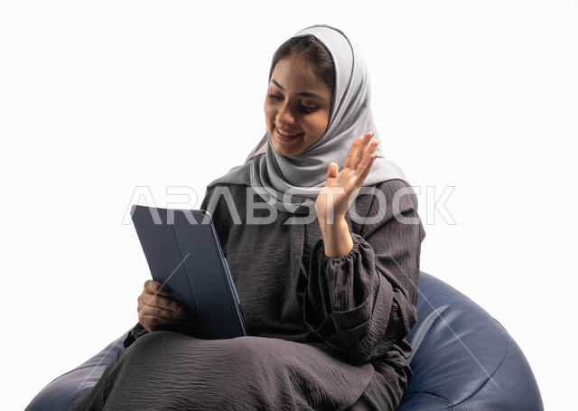 إستخدام اجهزة تقنية حديثة، إجراء مكالمات فيديو عبر الاونلاين، إيماءات وجه ويدين تدل على الترحيب, بورتريه لامرأة عربية سعودية خليجية تتجلس على كرسي مريح تستخدم جهاز التابلت ، التسوق الإلكتروني الآمن، تصفح الإنترنت وإنجاز المهام عن بعد, خلفية بيضاء 
