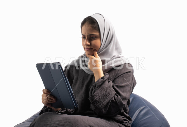 إيماءات وجه ويدين تعبر عن الحيرة بالتسوق و الشراء عبر الاونلاين, إستخدام اجهزة تقنية حديثة،  بورتريه لامرأة عربية سعودية خليجية تتجلس على كرسي مريح تستخدم جهاز التابلت ، التسوق الإلكتروني الآمن، تصفح الإنترنت وإنجاز المهام عن بعد، خلفية بيضاء
