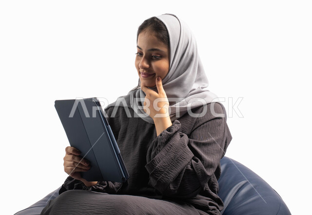 تسلية وترفيه, إستخدام اجهزة تقنية حديثة،  بورتريه لامرأة عربية سعودية خليجية تتجلس على كرسي مريح تستخدم جهاز التابلت ، التسوق الإلكتروني الآمن، تصفح الإنترنت وإنجاز المهام عن بعد ، الشراء والبيع عبر الاونلاين، خلفية بيضاء 