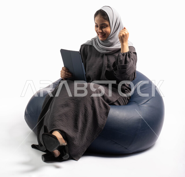 تصفح الإنترنت وإنجاز المهام عن بعد، إيماءات وجه ويدين تدل على الحماس, إستخدام اجهزة تقنية حديثة،  بورتريه لامرأة عربية سعودية خليجية تتجلس على كرسي مريح تستخدم جهاز التابلت ، التسوق الإلكتروني الآمن، الشراء والبيع عبر الاونلاين، خلفية بيضاء 
