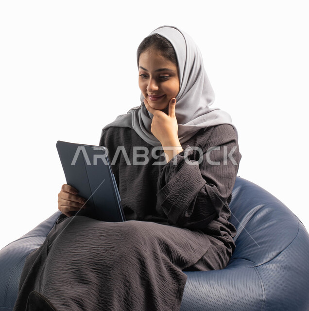 التسوق الإلكتروني الآمن، إيماءات وجه ويدين تعبر عن الاستمتاع بإستخدام اجهزة تقنية حديثة،  بورتريه لسيدة عربية سعودية خليجية تتجلس على كرسي مريح تستخدم جهاز التابلت ، تصفح الإنترنت وإنجاز المهام عن بعد ، الشراء والبيع عبر الاونلاين، خلفية بيضاء 