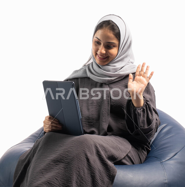 إجراء مكالمات فيديو عبر الاونلاين، إيماءات وجه ويدين تدل على الترحيب, إستخدام اجهزة تقنية حديثة،  بورتريه لامرأة عربية سعودية خليجية تتجلس على كرسي مريح تستخدم جهاز التابلت ، التسوق الإلكتروني الآمن، تصفح الإنترنت وإنجاز المهام عن بعد, خلفية بيضاء 