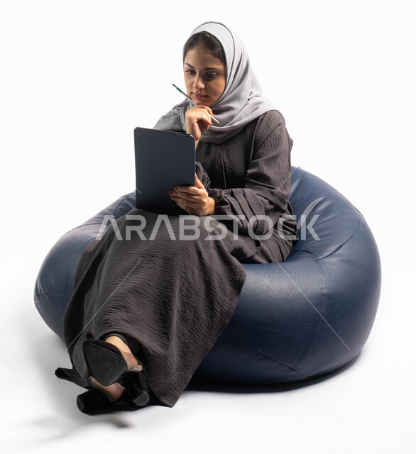 إيماءات وجه ويدين تعبر عن الحيرة, إستخدام اجهزة تقنية حديثة،  بورتريه لامرأة عربية سعودية خليجية تتجلس على كرسي مريح تستخدم جهاز التابلت ، التسوق الإلكتروني الآمن، تصفح الإنترنت وإنجاز المهام عن بعد ، الشراء والبيع عبر الاونلاين، خلفية بيضاء 