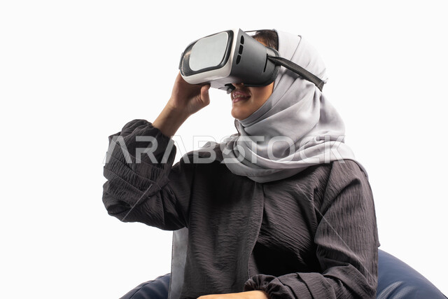 تقنية وألعاب حديثة, إيماءات تدل على الاستمتاع والتفاعل مع الألعاب الافتراضية، الاستمتاع بمشاهدة أفلام الواقع الافتراضي، بورتريه لإمرأة عربية خليجية سعودية تجلس على أريكة مريحة وترتدي نظارة الواقع الافتراضي ثلاثية الابعاد، خلفية بيضاء