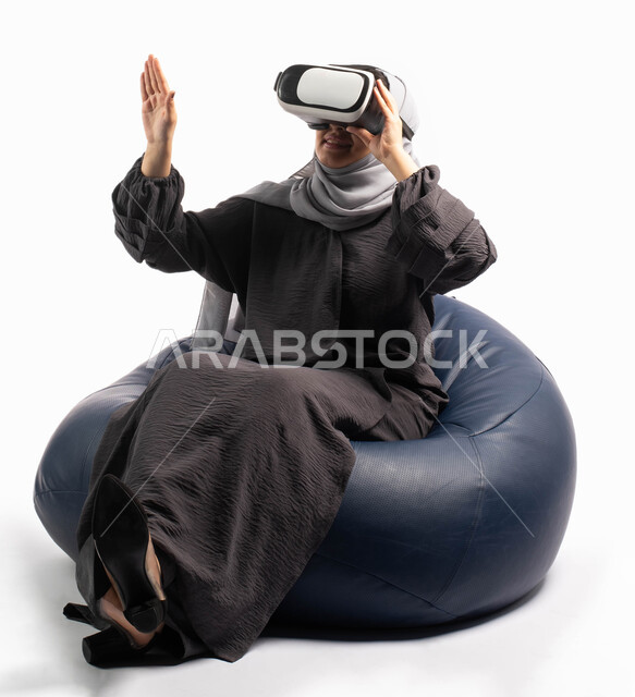 إيماءات تدل على الاستمتاع والتفاعل مع الألعاب الافتراضية، الاستمتاع بمشاهدة أفلام الواقع الافتراضي، بورتريه لإمرأة عربية خليجية سعودية تجلس على أريكة مريحة وترتدي نظارة الواقع الافتراضي ثلاثية الابعاد وتشير بيدها الى شيء ما، خلفية بيضاء