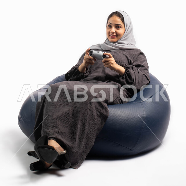 قضاء أوقات الفراغ باللعب بألعاب البلايستيشن الترفيهية ، إيماءات وتعابير وجه ويدين تدل على الاستمتاع باللعب, تقنيات حديثة مسلية, بورتريه لإمرأة عربية سعودية خليجية تجلس على كرسي مريح وتحمل بيدها عصا التحكم  و تلعب بألعاب الفيديو ، خلفية بيضاء