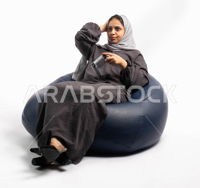 تقنيات حديثة مسلية, إيماءات وتعابير وجه ويدين تدل على الخسارة والهزيمة, قضاء أوقات الفراغ باللعب بألعاب البلايستيشن الترفيهية ، بورتريه لإمرأة عربية سعودية خليجية تجلس على كرسي مريح وتحمل بيدها عصا التحكم  و تلعب بألعاب الفيديو ، خلفية بيضاء