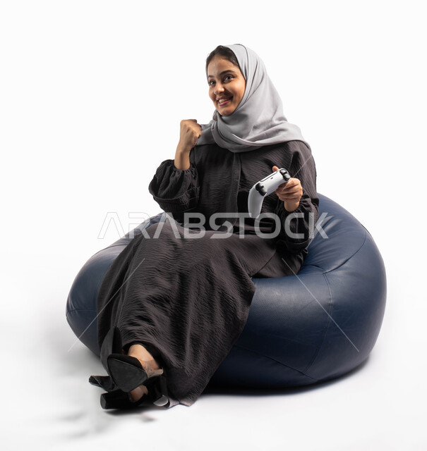 إيماءات وتعابير وجه ويدين تدل على الفوز و الانتصار, قضاء أوقات الفراغ باللعب بألعاب البلايستيشن الترفيهية ، تقنيات حديثة مسلية, بورتريه لإمرأة عربية سعودية خليجية تجلس على كرسي مريح وتحمل بيدها عصا التحكم  و تلعب بألعاب الفيديو ، خلفية بيضاء