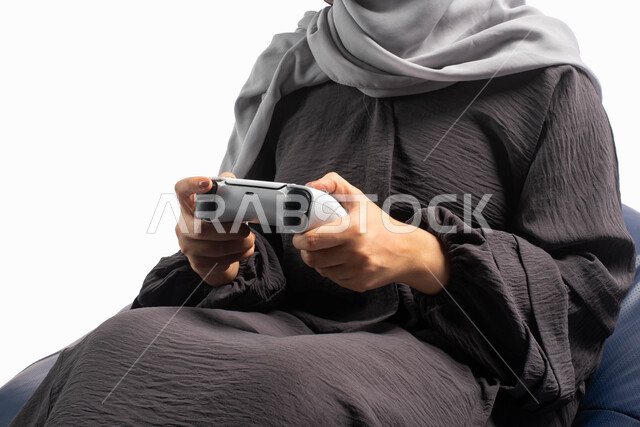 أنشطة ترفيهية ممتعة, قضاء أوقات الفراغ باللعب بألعاب البلايستيشن الترفيهية ، تقنيات حديثة مسلية, بورتريه صورة مقربة لإمرأة عربية سعودية خليجية تجلس على كرسي مريح وتحمل بيدها عصا التحكم  و تلعب بألعاب الفيديو ، خلفية بيضاء