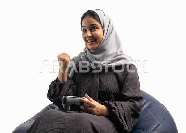 إيماءات وتعابير وجه ويدين تدل على الفوز و الانتصار, قضاء أوقات الفراغ باللعب بألعاب البلايستيشن الترفيهية ، تقنيات حديثة مسلية, بورتريه لإمرأة عربية سعودية خليجية تجلس على كرسي مريح وتحمل بيدها عصا التحكم  و تلعب بألعاب الفيديو ، خلفية بيضاء
