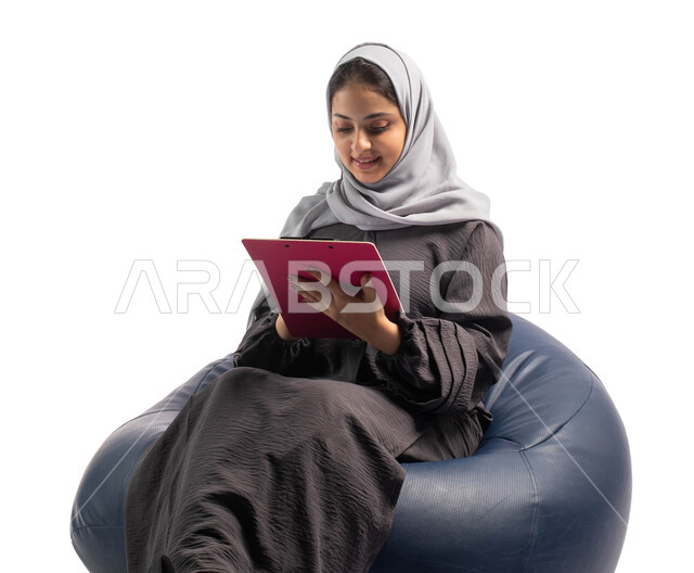 كتابة المهام والأهداف اليومية, إيماءات وتعابير وجه تعبر عن الاستمتاع بكتابة وتدوين الملاحظات الهامة، كتابة التقارير والابحاث على الورق، بورتريه لشابة عربية خليجية سعودية تجلس على كرسي مريح وتحمل بيدها أوراق وملفات خاصة بالعمل، خلفية بيضاء