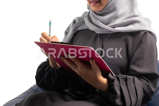  إيماءات وتعابير وجه تعبر عن الاستمتاع بكتابة وتدوين الملاحظات الهامة، كتابة التقارير والابحاث على الورق، بورتريه لشابة عربية خليجية سعودية تجلس على كرسي مريح وتحمل بيدها أوراق وملفات خاصة بالعمل، كتابة المهام والأهداف اليومية, خلفية بيضاء