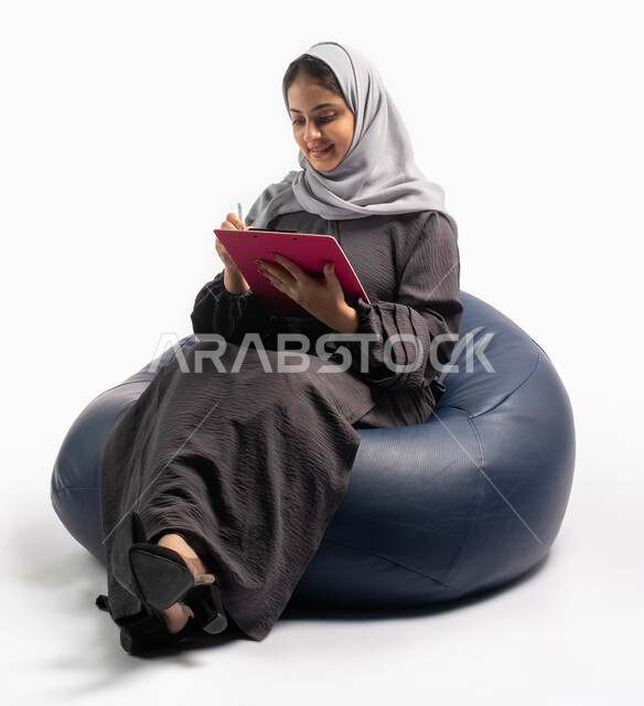 كتابة التقارير والابحاث على الورق، إيماءات وتعابير وجه تعبر عن الاستمتاع بكتابة وتدوين الملاحظات الهامة، بورتريه لشابة عربية خليجية سعودية تجلس على كرسي مريح وتحمل بيدها أوراق وملفات خاصة بالعمل، خلفية بيضاء