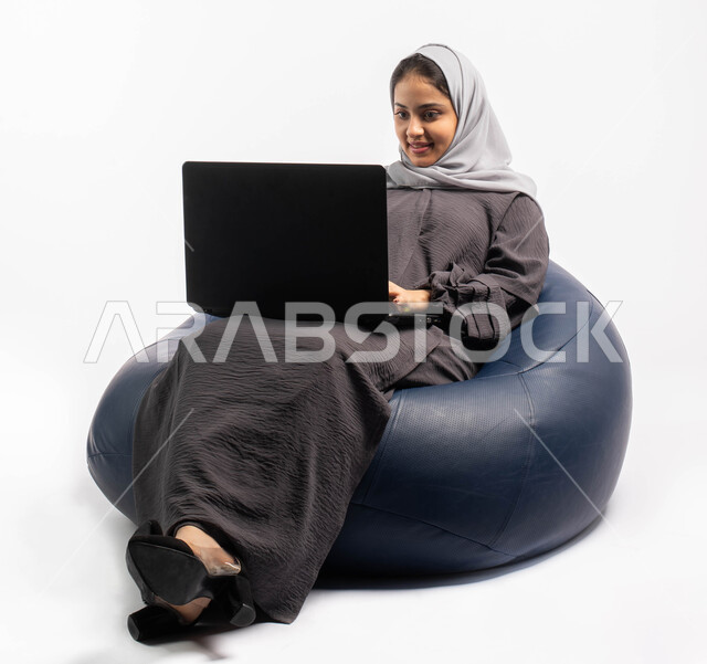 إجراء مكالمات فيديو عبر الإنترنت, إدارة المشاريع عن بعد, بورتريه صورة مقربة لإمرأة عربية سعودية خليجية تجلس على أريكة مريحة وتستخدم اللابتوب, انجاز مهام العمل عبر الحاسوب, اللعب على اللابتوب, قضاء وقت ممتع ومسلي, خلفيه بيضاء