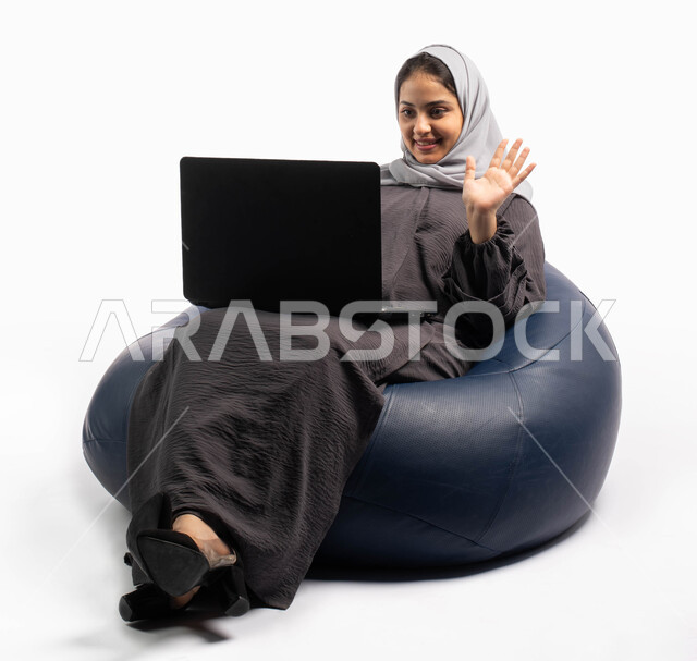 إيماءات وجه ويدين تدل على الترحيب, إدارة المشاريع عن بعد, بورتريه صورة مقربة لإمرأة عربية سعودية خليجية تجلس على كرسي مريح وتستخدم اللابتوب, انجاز مهام العمل عبر الحاسوب, إجراء مكالمات فيديو عبر الإنترنت, قضاء وقت ممتع, خلفيه بيضاء