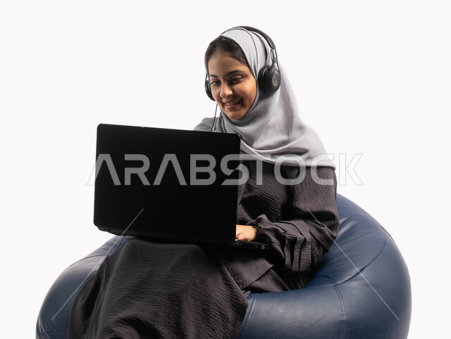 دردشة وتواصل عبر الانترنت، إيماءات وتعابير تدل على الاستمتاع باستخدام الحاسب المحمول, متابعة وتطوير الأعمال عن بعد، بورتريه لشابة عربية خليجية سعودية تجلس على كرسي مريح تستخدم الحاسوب المحمول و تضع سماعة الرأس، تسجيل الاغاني والصوتيات، خلفية بيضاء