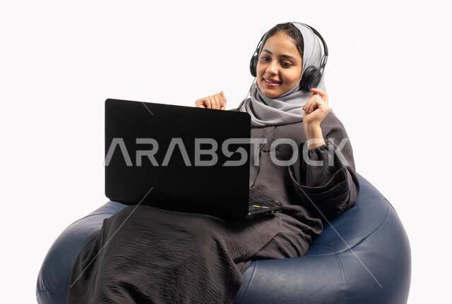 تسجيل الاغاني والصوتيات، إيماءات وتعابير تدل على الاستمتاع باستخدام الحاسب المحمول, متابعة وتطوير الأعمال عن بعد، بورتريه لشابة عربية خليجية سعودية تجلس على كرسي مريح تستخدم الحاسوب المحمول و تضع سماعة الرأس، دردشة وتواصل عبر الانترنت، خلفية بيضاء
