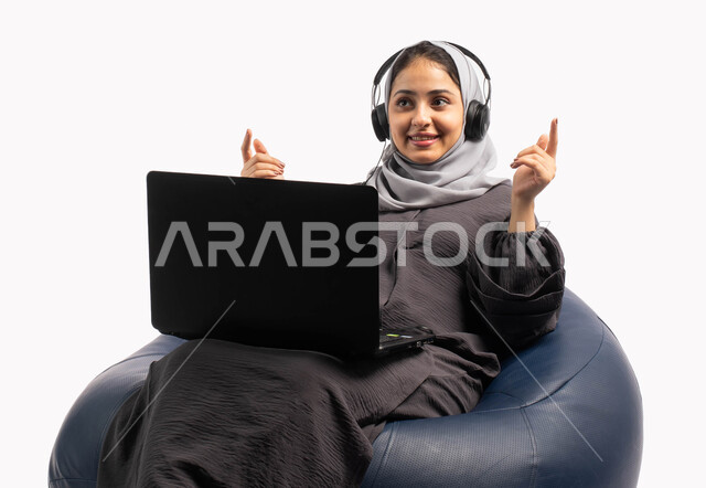 إيماءات وتعابير تدل على الاستمتاع باستخدام الحاسب المحمول, متابعة وتطوير الأعمال عن بعد، بورتريه لشابة عربية خليجية سعودية تجلس على كرسي مريح تستخدم الحاسوب المحمول و تضع سماعة الرأس، تسجيل الاغاني والصوتيات، دردشة وتواصل عبر الانترنت، خلفية بيضاء