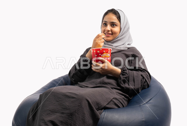 Entertaining and entertaining by spending fun and entertaining times, enjoying watching movies, facial and hand gestures indicating enjoyment of watching a movie, a portrait of a young Saudi Arabian Gulf woman sitting on a comfortable sofa and holding a b