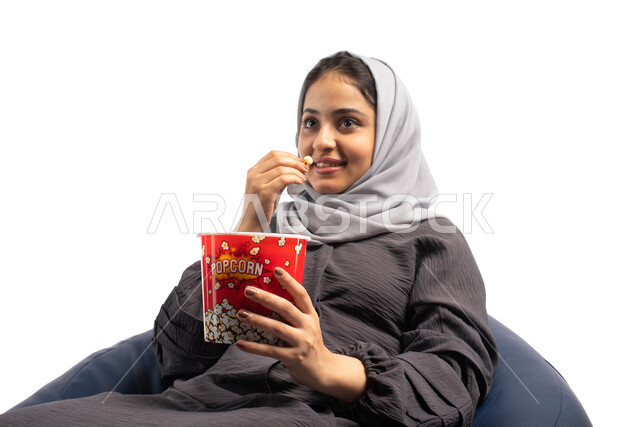 التسلية والترفيه بقضاء أوقات ممتعة ومسلية، الاستمتاع بمشاهدة الأفلام، ايماءات وجه ويدين تدل على الاستمتاع بمتابعة فيلم سينمائي،  بورتريه لشابة عربية خليجية سعودية تجلس على أريكة مريحة وتحمل بيدها علبة الفوشار، خلفية بيضاء