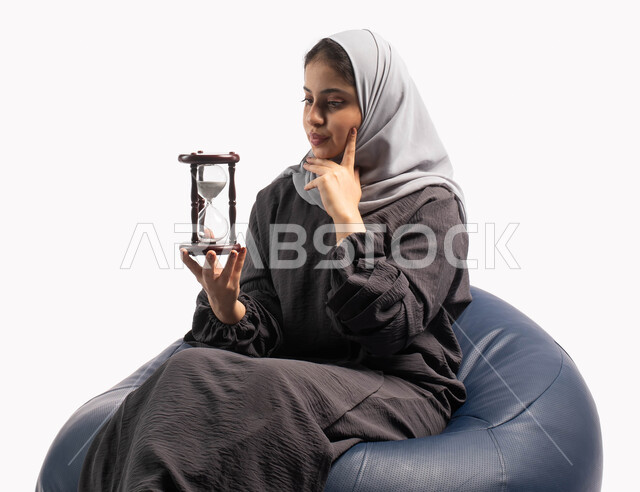 Deep mental thought, thinking about the past and the future, the concept of reflection and focus on time, a portrait of a Saudi Gulf Arab woman sitting on a comfortable chair and holding an hourglass in her hand and placing her hand on her cheek, remember