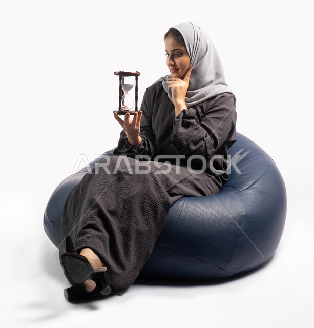 التحليل واتخاذ القرارات, التفكير في الماضي والمستقبل, مفهوم التمعن والتركيز بالوقت, بورتريه لإمرأة سعودية خليجية عربية تجلس على كرسي مريح وتمسك بيدها ساعة رملية وتضع يدها على خدها, تذكر ذكريات ومواقف جميلة, الفكر الذهني العميق, خلفية بيضاء