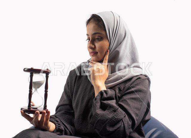 مفهوم التمعن والتركيز بالوقت, التفكير في الماضي والمستقبل, بورتريه لإمرأة سعودية خليجية عربية تجلس على كرسي مريح وتمسك بيدها ساعة رملية وتضع يدها على خدها, تذكر ذكريات ومواقف جميلة, الفكر الذهني العميق, التحليل واتخاذ القرارات, خلفية بيضاء
