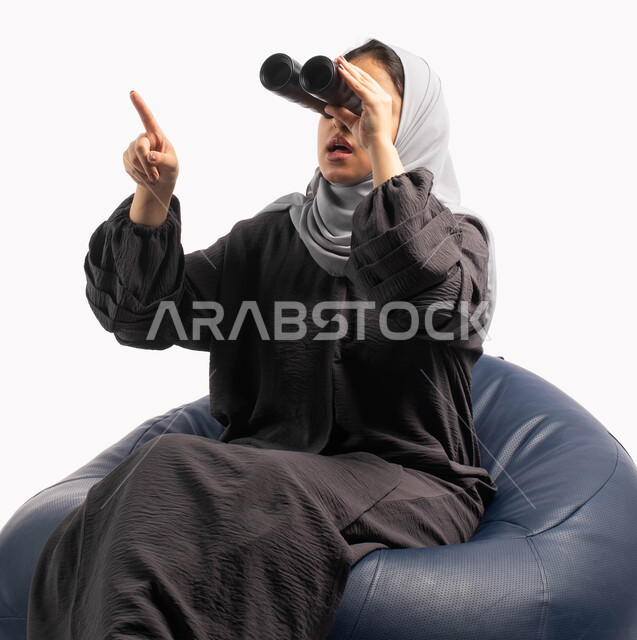 رؤية مستقبلية لدى اليافعين، حب الاستكشاف والمغامرة، إيماءات وتعابير وجه تدل على الدهشة و الاستغراب, التخطيط المستقبلي للأهداف و الطموحات، بورتريه لإمرأة عربية خليجية سعودية تجلس على كرسي مريح وتنظر من خلال منظار الرؤية وتشير بيدها الى شيء ما, خلفية بيضاء