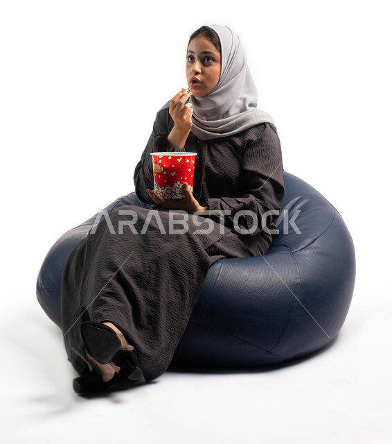 الاستمتاع بمشاهدة الأفلام، ايماءات وجه ويدين تدل على الدهشة والاستغراب، بورتريه لإمرأة عربية خليجية سعودية تجلس على كرسي مريح وتحمل بيدها علبة الفوشار، مشاهدة فيلم سينمائي، التسليه والترفيه بقضاء أوقات ممتعة ومسلية، خلفية بيضاء