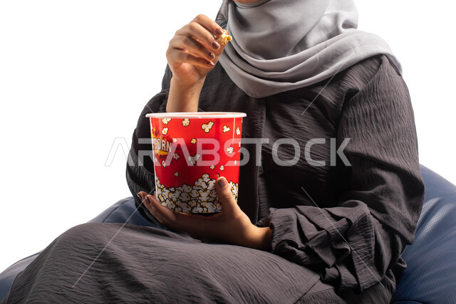 أنشطة ترفيهية ممتعة , بورتريه لإمرأة عربية خليجية سعودية تجلس على كرسي مريح وتحمل بيدها علبة الفوشار، الاستمتاع بمشاهدة الأفلام، مشاهدة فيلم سينمائي، التسليه والترفيه بقضاء أوقات ممتعة ومسلية، خلفية بيضاء