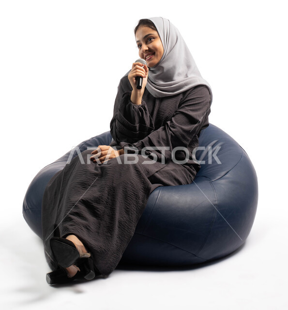 ايماءات وتعابير وجه و يدين تعبر عن الاستمتاع بالغناء، فن إلقاء الخطابات و المحاضرات، التدريب الصوتي وتقديم البرامج الصوتية، بورتريه لسيدة عربية خليجية سعودية تجلس على أريكة مريحة وتحمل بيدها الميكروفون، خلفية بيضاء
