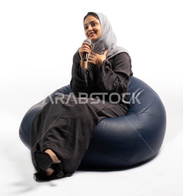  ايماءات وتعابير وجه و يدين تعبر عن الاستمتاع بالغناء، فن إلقاء الخطابات و المحاضرات، التدريب الصوتي وتقديم البرامج الصوتية، بورتريه لسيدة عربية خليجية سعودية تجلس على أريكة مريحة وتحمل بيدها الميكروفون، خلفية بيضاء