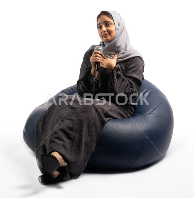 التدريب الصوتي وتقديم البرامج الصوتية، فن إلقاء الخطابات والندوات، بورتريه لشابة عربية خليجية سعودية تجلس على كرسي مريح وتمسك بيدها الميكروفون، ايماءات وتعابير وجه و يدين تعبر عن الاستمتاع والطرب، خلفية بيضاء