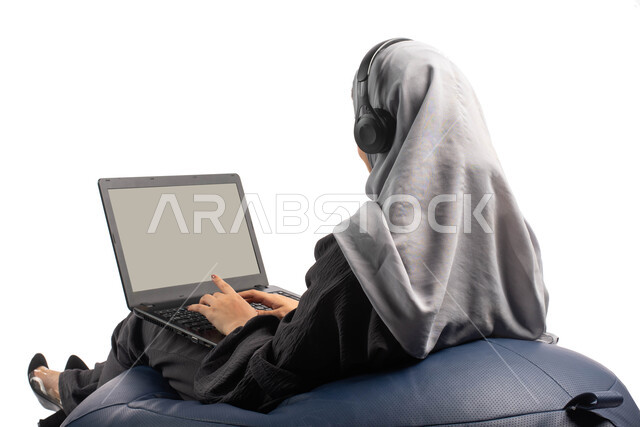 Follow-up and business development, completing remote work tasks, a portrait from behind of a Saudi Arabian Gulf woman sitting on a comfortable chair using a laptop and wearing a headset, recording songs and audios, chatting and communicating via the Inte