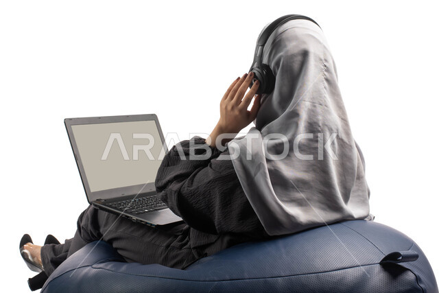 انجاز مهام العمل عن بعد، متابعة وتطوير الأعمال، بورتريه من الخلف لشابة عربية خليجية سعودية تجلس على كرسي مريح تستخدم الحاسوب المحمول و تضع سماعة الرأس، تسجيل الاغاني والصوتيات، دردشة وتواصل عبر الانترنت، خلفية بيضاء