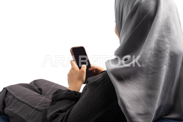  استخدام البرامج والتطبيقات الالكترونية، قضاء وقت ممتع, بورتريه من الخلف لإمرأة عربية خليجية سعودية تجلس على كرسي مريح وتقوم بتصفح الهاتف المحمول ، التسوق الالكتروني عبر الانترنت، تصفح المواقع الالكترونية والتواصل الاجتماعي، خلفية بيضاء