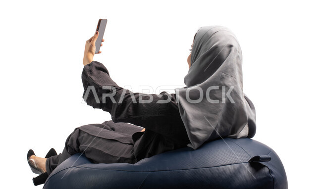 اجراء مكالمة فيديو من خلال الهاتف الجوال، التواصل والتحدث مع الأهل والأصدقاء, بورتريه من الخلف لإمرأة عربية خليجية سعودية تجلس على كرسي مريح وتمسك بيدها الهاتف المحمول، خلفية بيضاء