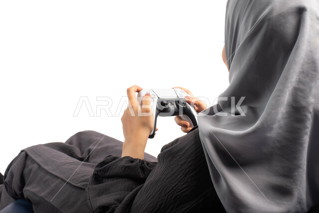 قضاء أوقات الفراغ باللعب بألعاب البلايستيشن الترفيهية ، تقنيات حديثة مسلية, بورتريه من الخلف لإمرأة عربية سعودية خليجية تجلس على كرسي مريح وتحمل بيدها عصا التحكم  و تلعب بألعاب الفيديو ، خلفية بيضاء