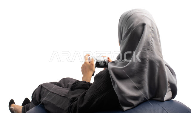 تقنيات حديثة مسلية, بورتريه من الخلف لإمرأة عربية سعودية خليجية تجلس على كرسي مريح وتحمل بيدها عصا التحكم  و تلعب بألعاب الفيديو ، قضاء أوقات الفراغ باللعب بألعاب البلايستيشن الترفيهية ، خلفية بيضاء